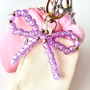 May include: A beaded bow keychain featuring translucent purple faceted beads accented with small metallic gold spacers. The bow is attached to a silver and gold-toned metal lobster clasp and ring hardware, designed to be clipped onto bags or accessories.