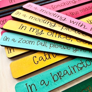 May include: A set of colorful wooden signs with different phrases for a teacher's classroom. The phrases include "In a data/PLC meeting", "In a meeting - please give me a few minutes", "In my office, on a Zoom call, please give me a few minutes", "Gather", "In a brainstorming session", and "Classroom".