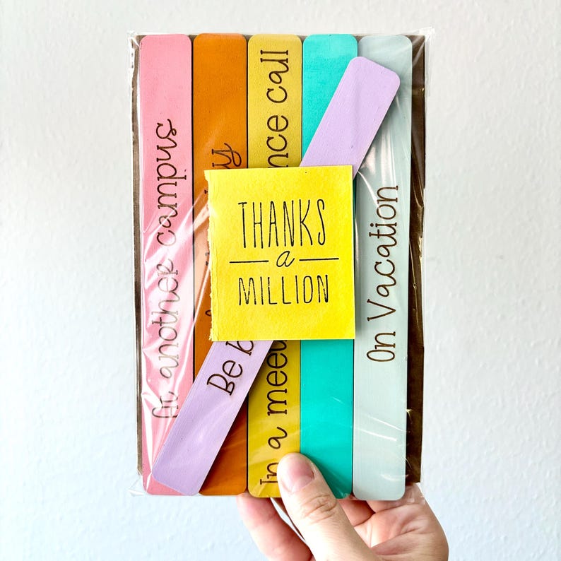 May include: Set of six colorful wooden bookmarks with different phrases, including "Thanks a Million", "On Vacation", "Be Kind", "In a meeting", "Once call", and "In another campus".