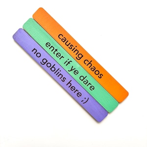 May include: Three wooden sticks painted in purple, green, and orange with the text "causing chaos enter if ye dare no goblins here ;)" written on them.