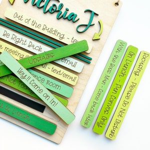 May include: A wooden sign with the name "Victoria J" written in green cursive. The sign has a sliding system with different colored wooden blocks that say "Out of the Building - text/email", "Be Right Back - Upstairs", "a meeting - once room", "Hey!! Come on in! - Not Today", "With the Boss Man - Come back later", "On Lunch - Fun Convos Only", and "Focusing - Please Do Not Disturb".