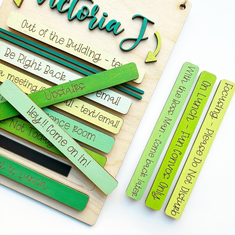May include: A wooden sign with the name "Victoria J" written in green cursive. The sign has a sliding system with different colored wooden blocks that say "Out of the Building - text/email", "Be Right Back - Upstairs", "a meeting - once room", "Hey!! Come on in! - Not Today", "With the Boss Man - Come back later", "On Lunch - Fun Convos Only", and "Focusing - Please Do Not Disturb".