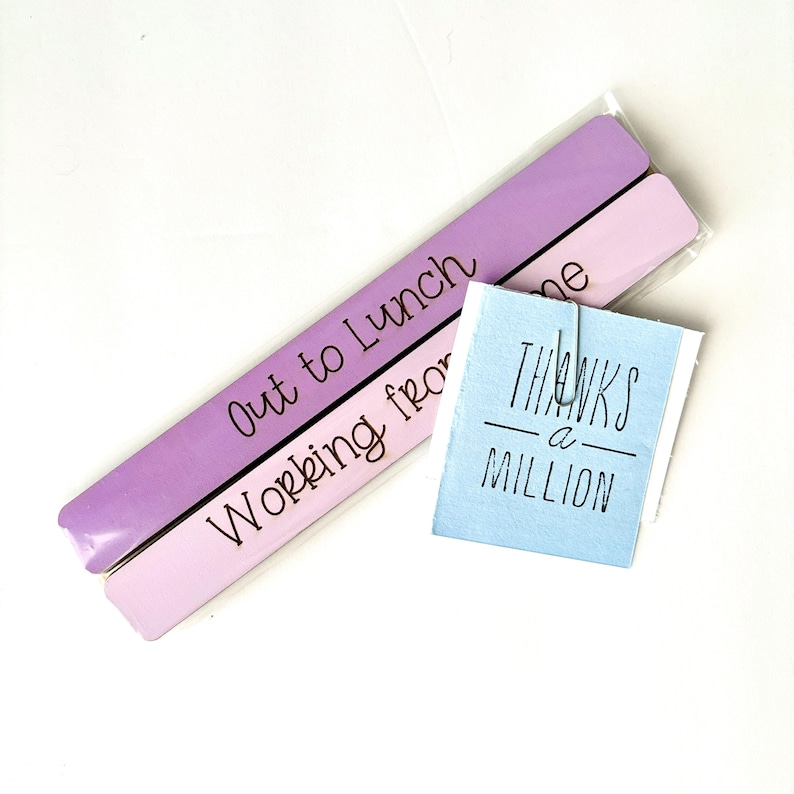 May include: Two purple and pink wooden signs with the text "Out to Lunch" and "Working from home". A blue card with the text "Thanks a Million" is attached to the signs with a silver paper clip.