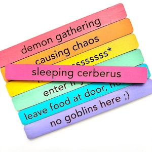 May include: A set of six colorful wooden sticks with different phrases written on them. The phrases are: "demon gathering", "causing chaos", "sleeping cerberus", "enter", "leave food at door, ho", and "no goblins here ;)".
