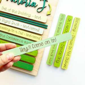 May include: A wooden sign with the text "Victoria J" at the top. The sign has a series of green and yellow wooden slats with text on each slat. The text on the slats includes "Out of the Building - text", "Right Back - text/email", "Meeting/On a Call - text/email", "The Conference Room", "Focusing - Please Do Not Disturb", "On Lunch", "With the Boss - Convos Only", and "Hey!! Come on in!"