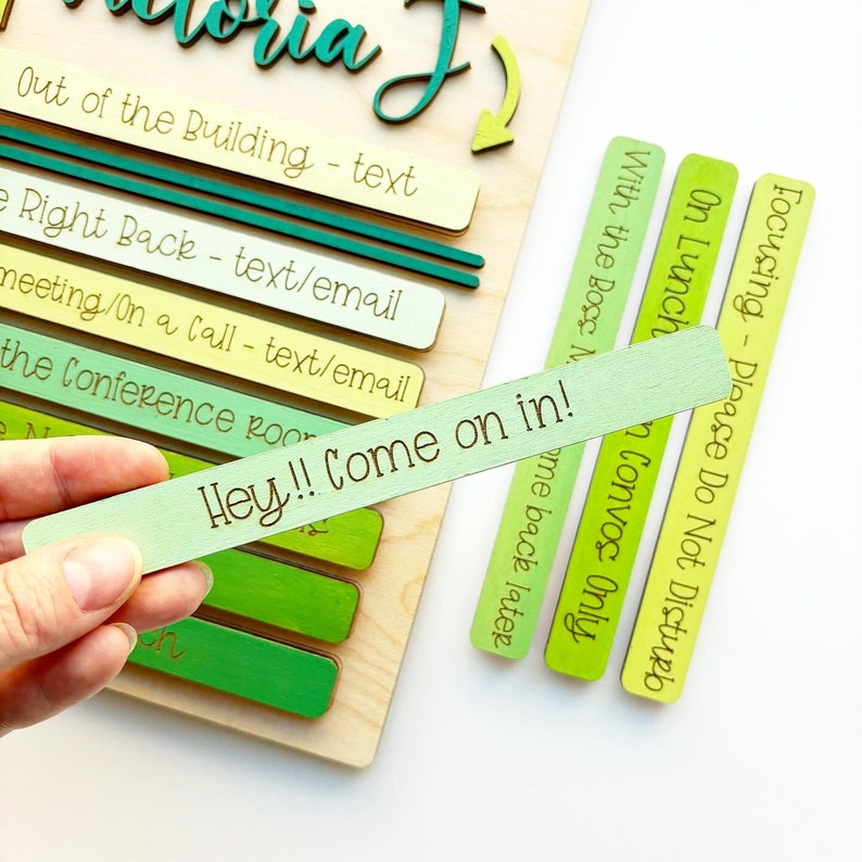 May include: A wooden sign with the text "Victoria J" at the top. The sign has a series of green and yellow wooden slats with text on each slat. The text on the slats includes "Out of the Building - text", "Right Back - text/email", "Meeting/On a Call - text/email", "The Conference Room", "Focusing - Please Do Not Disturb", "On Lunch", "With the Boss - Convos Only", and "Hey!! Come on in!"