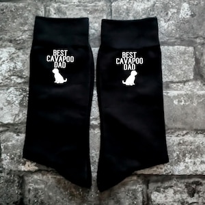 May include: A pair of black crew socks featuring the white text BEST CAVAPOO DAD printed above a white silhouette of a small dog. The socks have a solid black fabric construction with a ribbed cuff at the top and a simple graphic design.