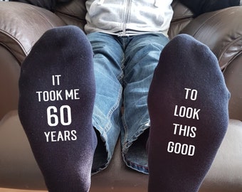May include: A pair of dark navy blue socks featuring white text printed on the soles. The left sock reads IT TOOK ME 60 YEARS and the right sock reads TO LOOK THIS GOOD. The socks are worn on feet positioned forward, showing the printed soles clearly.