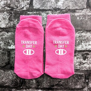 May include: A pair of bright pink ankle socks featuring the white text TRANSFER DAY, two small gold hearts, and a white oval graphic with two vertical lines inside, centred on the front of each sock.