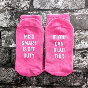 May include: A pair of pink socks with white text. One sock says "Miss Smart is off duty" and the other sock says "If you can read this".