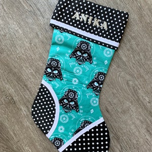Puede incluir: Una media de Navidad de color azul verdoso y negro con un patrón de lunares blancos. La media presenta un patrón repetido de la cara de Darth Vader con un diseño de calavera de azúcar. La media está personalizada con el nombre "Anika" en bordado blanco.
