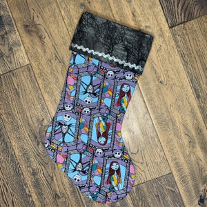 May include: A purple and blue Christmas stocking with a stained glass pattern featuring characters from the Nightmare Before Christmas. The stocking has a black and white fabric cuff with a silver lace trim.
