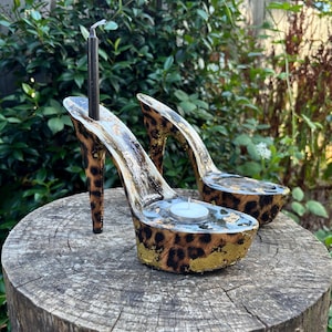 May include: A pair of decorative candle holders shaped like high-heeled shoes. The shoes have a leopard print design with gold accents. One shoe holds a black candle, the other a white tea light candle. The candle holders are displayed on a tree stump.
