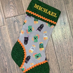 May include: A green and gray Christmas stocking with an orange trim. The stocking has a Minecraft theme with images of Creeper and Enderman characters. The stocking is personalized with the name "MICHAEL" in gold glitter.