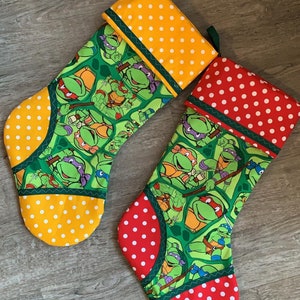 Puede incluir: Dos calcetines de Navidad con temática de las Tortugas Ninja. Un calcetín es rojo con lunares blancos y el otro es amarillo con lunares blancos. Ambos calcetines tienen una tela verde con imágenes de las Tortugas Ninja.