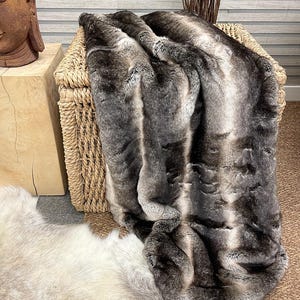 May include: A soft, gray and white faux fur throw blanket with a striped pattern. The blanket is draped over a wicker basket.