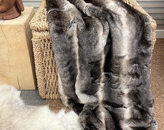 May include: A soft, grey and white faux fur throw blanket with a striped pattern. The blanket is draped over a wicker basket.