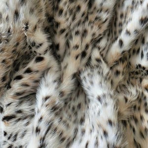 May include: A faux fur fabric featuring a dense, soft texture with a spotted animal print pattern. The material displays a color palette of cream, white, and tan base tones, accented by numerous small, irregular black and dark brown spots.