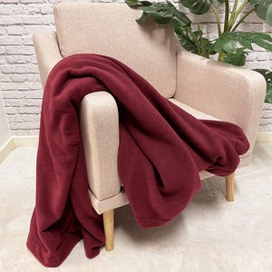 Puede incluir: Una manta de forro polar burdeos drapeada sobre un sillón beige con patas de madera. La manta es suave y acogedora, perfecta para acurrucarse en un día frío.