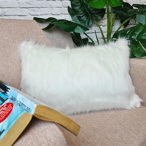May include: A rectangular white faux fur pillow rests on a pink sofa. The pillow has a fluffy texture and is positioned near an open book with a blue cover. Green leafy plants are in the background.