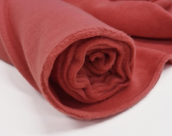 May include: Rolled-up, soft, coral-colored fleece fabric. The fabric is a solid color with a slightly textured surface. The edges of the fleece are visible, showing the thickness and texture of the material. The fleece is tightly rolled, creating a spiral pattern.