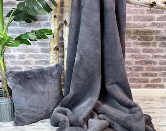 May include: A soft, grey faux fur throw blanket and matching cushion. The blanket is draped over a branch and the cushion is lying on a wooden floor.