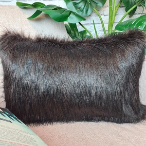 May include: A black faux fur throw pillow with a soft, plush texture. The pillow is sitting on a light brown couch.