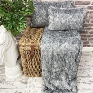 May include: A gray faux fur throw blanket and two matching throw pillows are displayed on a wicker storage bin. The throw blanket and pillows are soft and fluffy, and the wicker bin is a natural woven material.