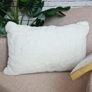 Puede incluir: Un cojín de sofá blanco de piel sintética con textura esponjosa. El cojín está colocado en un sofá marrón claro.