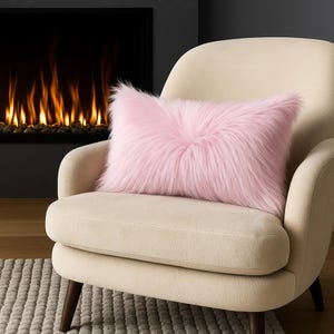 May include: A rectangular, light pink faux fur pillow rests on a cream-colored armchair. The pillow has a fluffy, textured appearance. The chair has a curved back and wooden legs. A fireplace with a dark surround is in the background.
