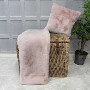 May include: A pink faux fur throw blanket and cushion set. The throw is draped over a wicker storage trunk. The cushion is sitting on top of the trunk.