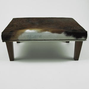 May include: A brown and white cowhide ottoman with wooden legs. The ottoman has a decorative nailhead trim around the edge.