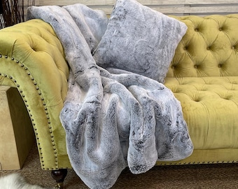 May include: A grey faux fur throw blanket and cushion are draped over a yellow tufted sofa. The throw is soft and fluffy, and the cushion is a great accent piece.