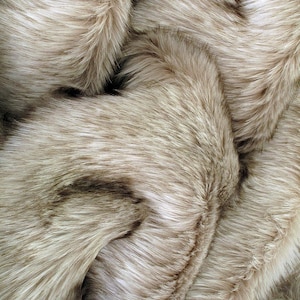 Può includere: Primo piano di un tessuto in finta pelliccia morbido nei toni del beige e del marrone. La pelliccia ha fibre lunghe e morbide con un aspetto naturale e strutturato. Il tessuto è piegato, creando ombre e profondità.