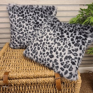 May include: Two gray and white faux fur throw pillows with a leopard print pattern. The pillows are sitting on a woven wicker basket.