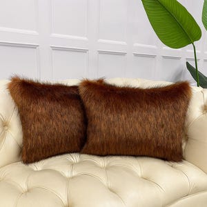 May include: Two rectangular brown faux fur throw pillows. The pillows have a shaggy texture and are placed on a cream-colored tufted sofa. The pillows add a touch of luxury and warmth to the home decor.