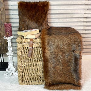 May include: A brown faux fur throw blanket and pillow set. The throw is draped over a wicker storage basket. The pillow is sitting on top of the basket.