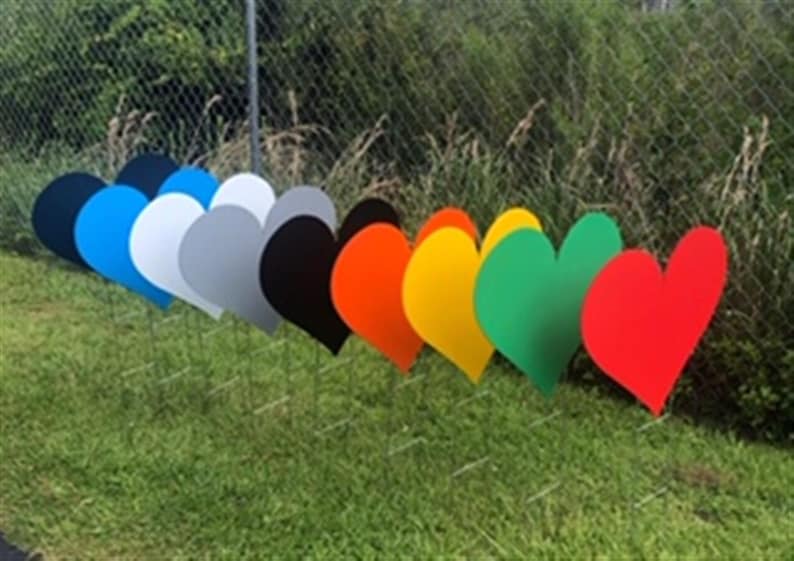 Pu&ograve; includere: Una fila di decorazioni da giardino a forma di cuore colorate in vari colori: blu, bianco, grigio, nero, arancione, giallo, verde e rosso. I cuori sono posizionati nell'erba vicino a una recinzione a catena.