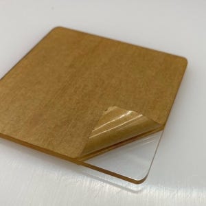 May include: A square piece of clear acrylic with a brown backing. The backing has a protective paper covering that is partially peeled back.