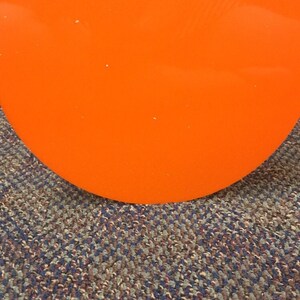 May include: Bright orange, circular object with the text "SIBE POLYMERS" in black. The object is set against a textured carpet with a mix of blue, brown, and gray fibers. The image is well-lit.