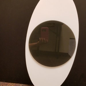 May include: An oval-shaped white mirror with a circular dark gray mirror in the center. The mirror reflects a person holding a pink phone. The background is dark brown.