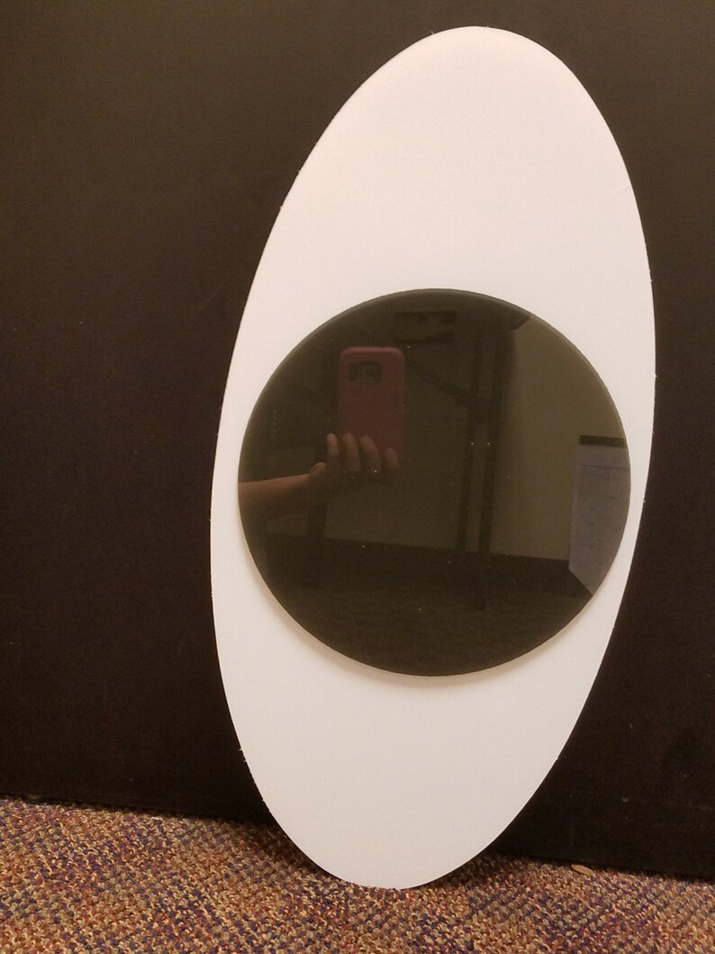 Op de afbeelding: Een ovaalvormige witte spiegel met een cirkelvormige donkergrijze spiegel in het midden. De spiegel reflecteert een persoon die een roze telefoon vasthoudt. De achtergrond is donkerbruin.