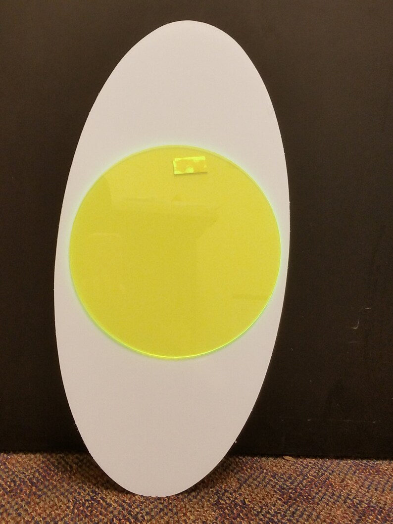 May include: An egg-shaped object, white with a central yellow circle. A small, rectangular yellow piece is attached to the yellow circle. The object is set against a dark background.