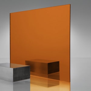 May include: A large sheet of orange acrylic glass, standing upright on a white surface. A smaller, rectangular, silver metal block sits in front of the glass.