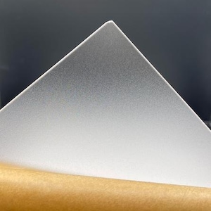 May include: A frosted, triangular sheet of translucent material, possibly acrylic or plastic, with a textured surface. The sheet is partially peeled back, revealing a brown paper backing. The background is a dark gray.