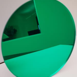 May include: A circular, emerald green object with a reflective surface. The object is positioned at an angle, revealing a glimpse of a room's interior. The edges are smooth and the color is consistent throughout.