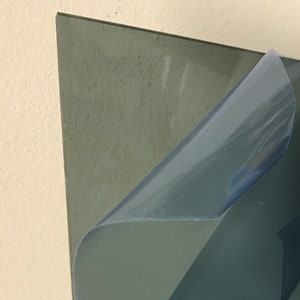 May include: A corner view of a gray, rectangular sheet with a protective blue film being peeled back. The sheet has a textured surface and a dark gray color. The protective film is partially peeled, revealing the sheet's surface.