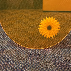 May include: A yellow daisy flower on a yellow, round, translucent surface. The surface is on a patterned carpet with brown, blue, and gray colors.