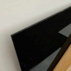 May include: A close-up of a black, rectangular object with a glossy surface. The object is positioned at an angle, with a portion of a tan-colored surface visible in the lower right corner. The background is a plain white surface.