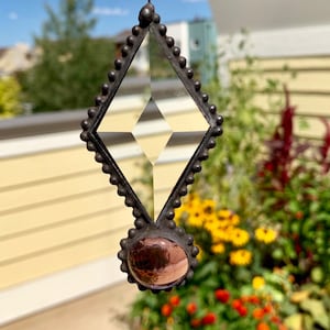 May include: A diamond-shaped stained glass suncatcher with a clear, geometric design. The suncatcher has a dark metal frame with a beaded edge and a round, purple-toned glass accent at the bottom. It hangs from a metal ball chain.